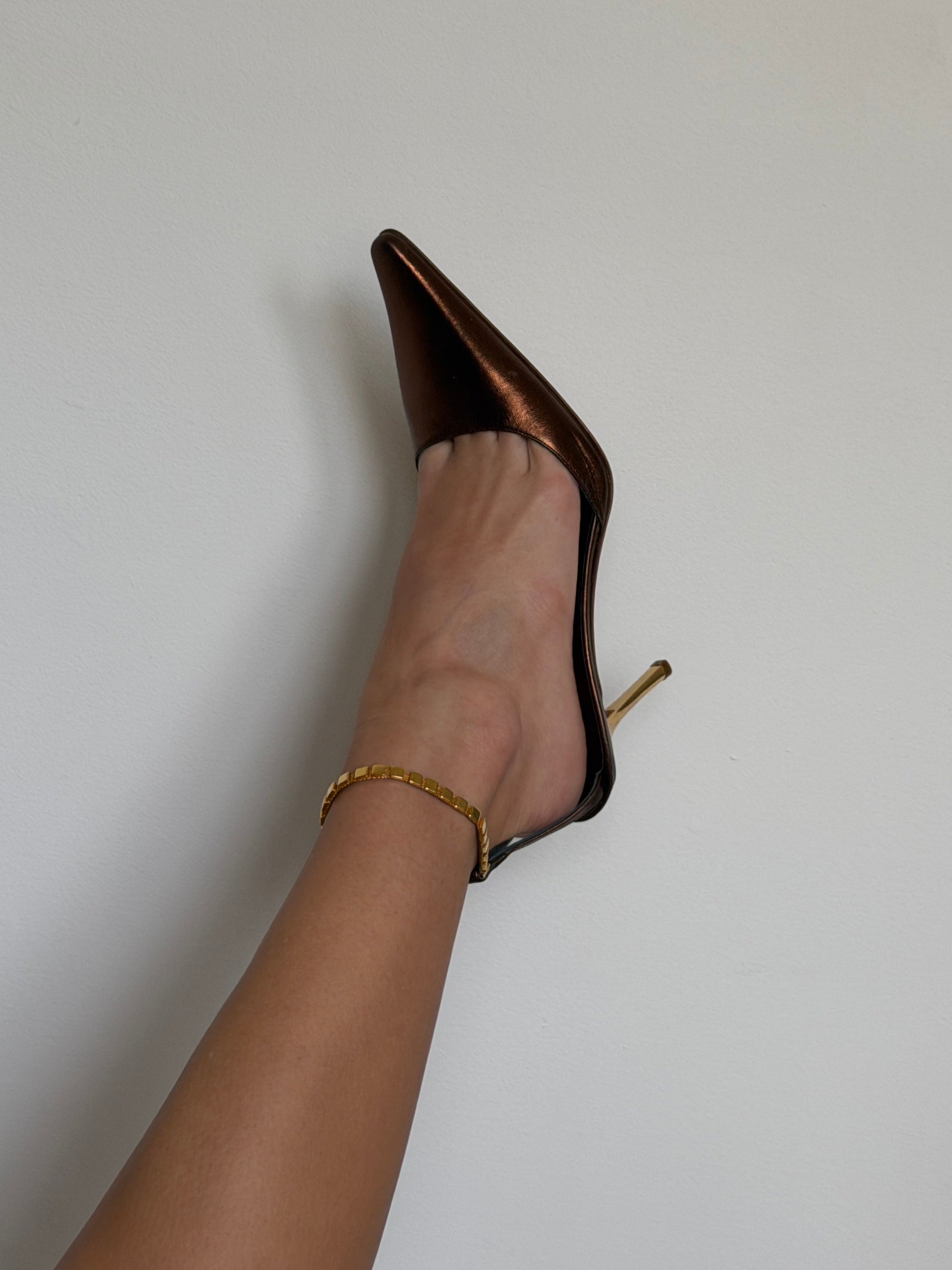 Brown high-heeled shoes worn with a gold ankle bracelet on a plain background