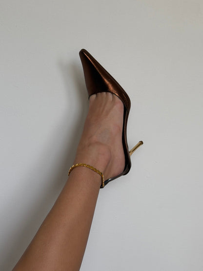 Brown high-heeled shoes worn with a gold ankle bracelet on a plain background