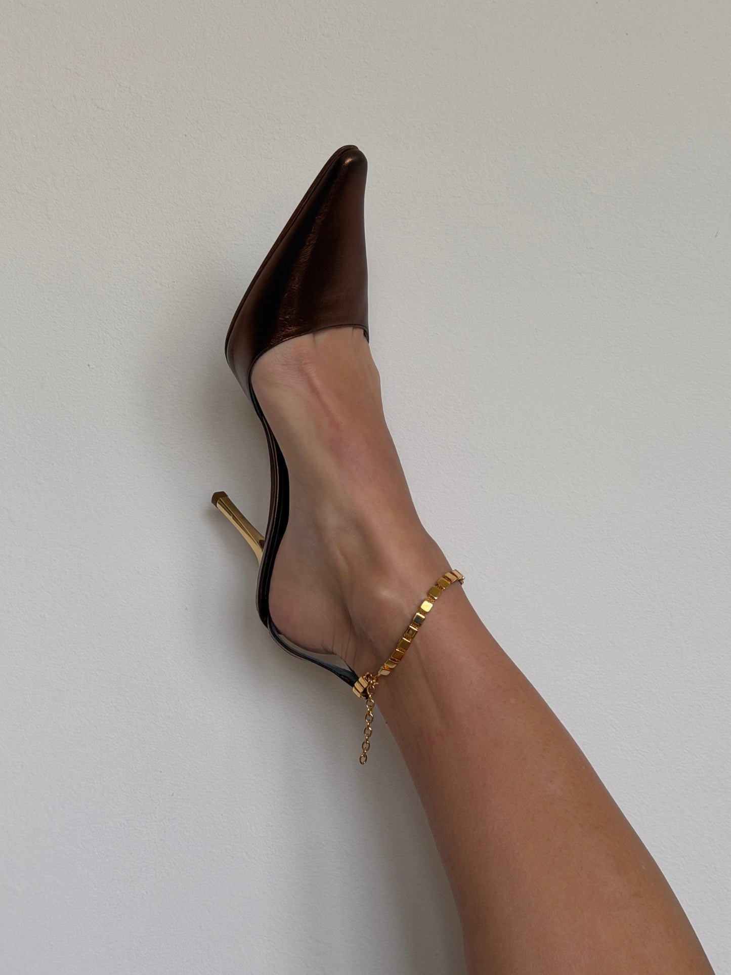 Brown high-heeled shoe worn with a gold ankle strap on a light gray background