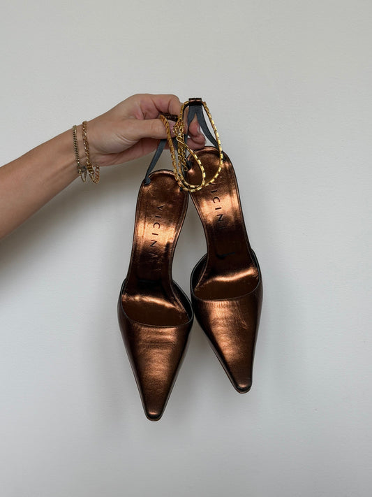 Bronze high-heeled shoes held by a hand against a plain background
