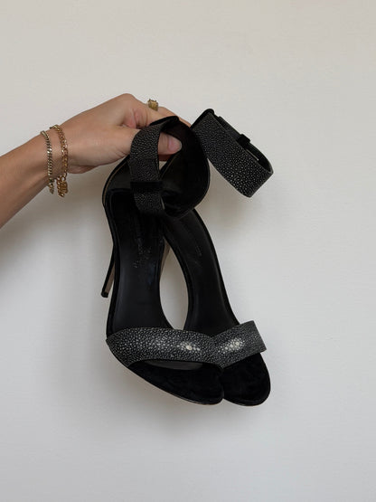 Black high-heeled shoes with glittery detail held by a hand on a light gray background