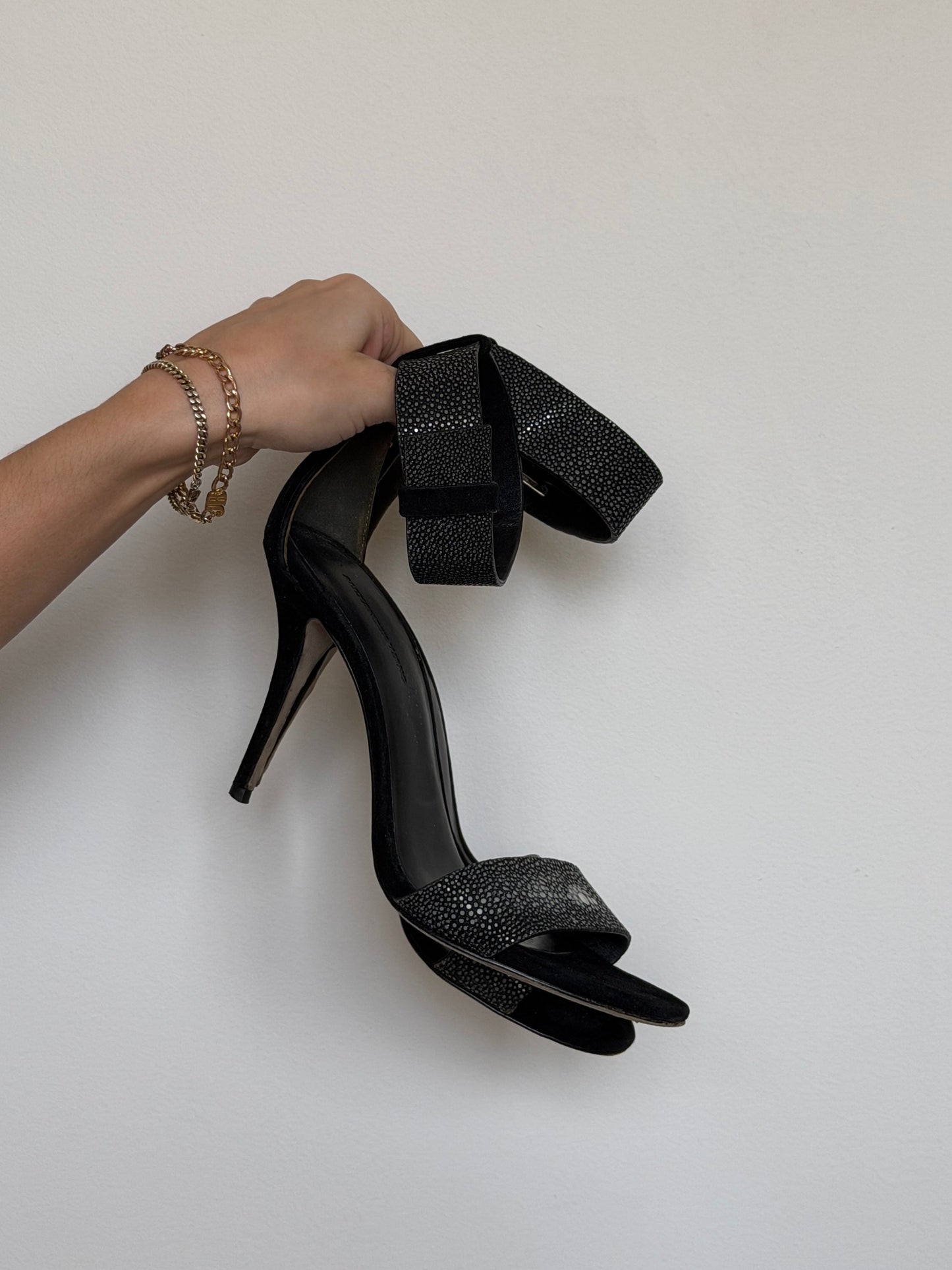 Black high-heeled shoes with a bow held by a hand on a light gray background