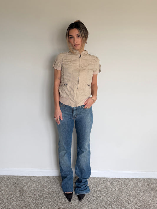 Person wearing a beige short-sleeve shirt and blue jeans against a plain wall.