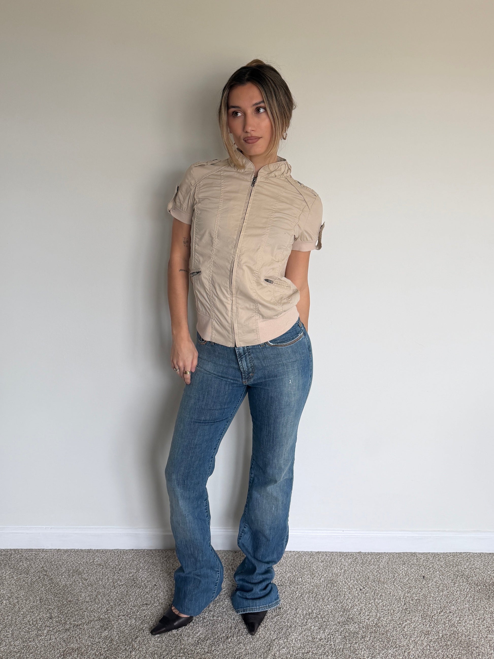 Person wearing a beige short-sleeve shirt and blue jeans against a plain wall.