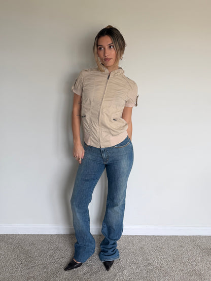 Person wearing a beige short-sleeve shirt and blue jeans against a plain wall.