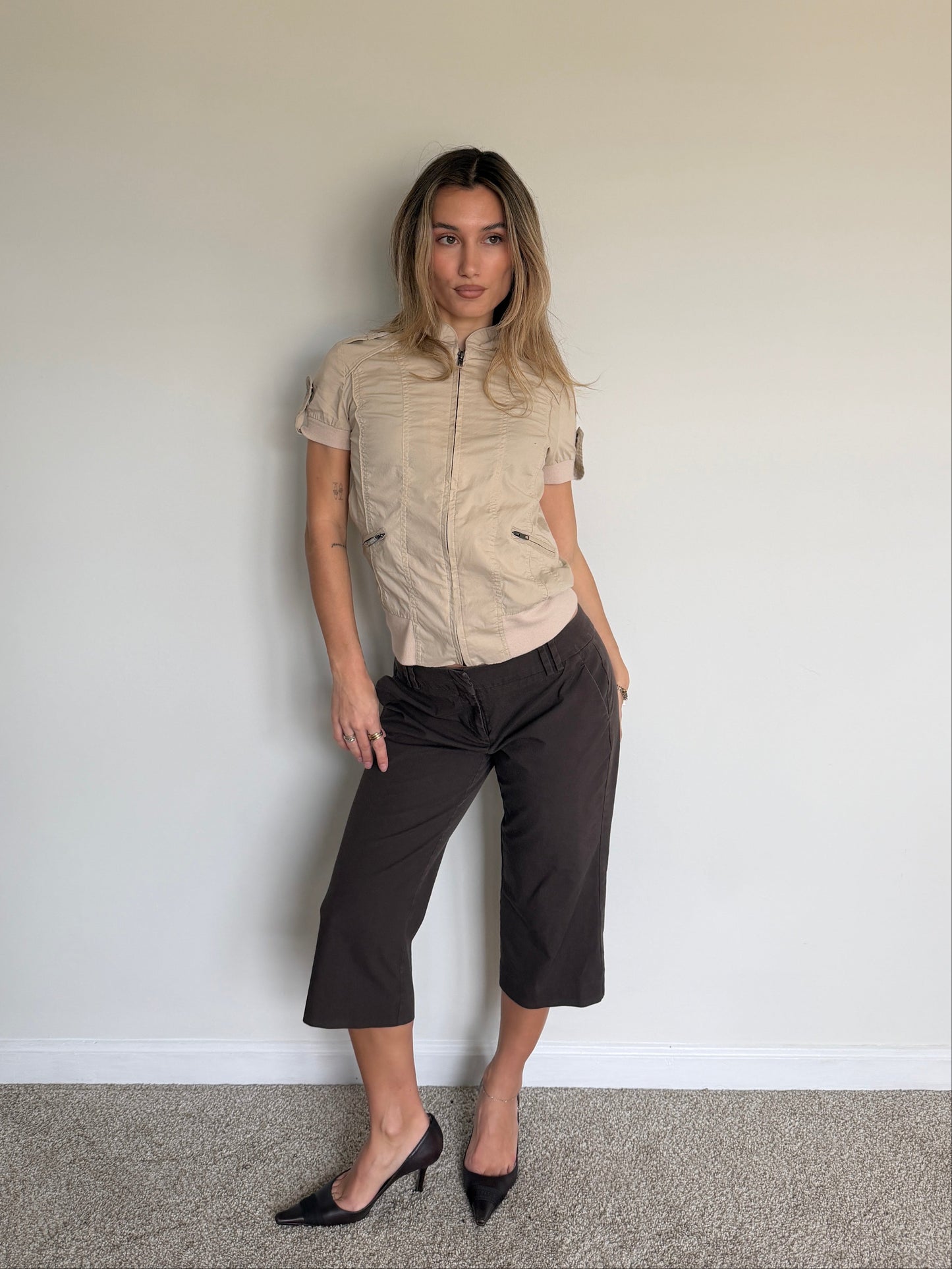 Woman wearing a beige short-sleeve shirt and black pants against a plain wall.