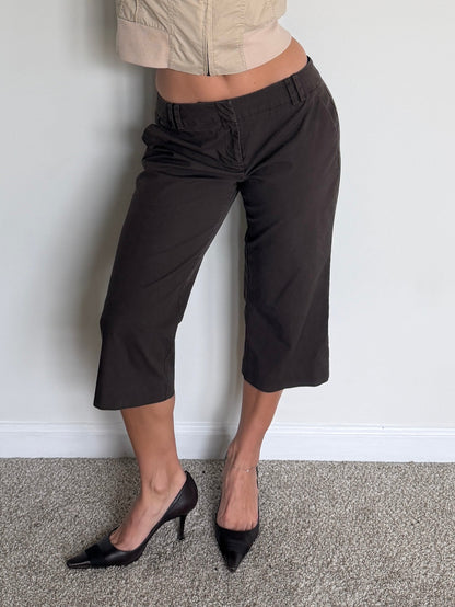 Person wearing a beige sleeveless jacket and black pants against a plain wall.