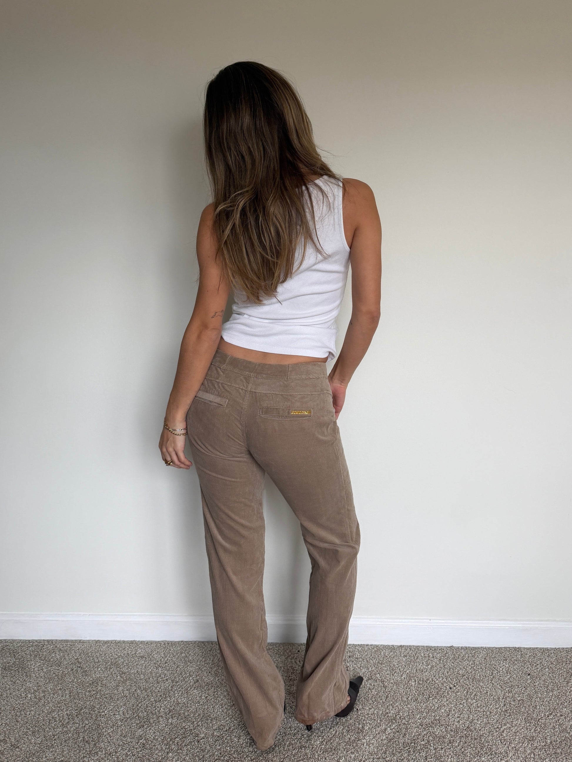 Person wearing a white tank top and brown pants against a plain wall.