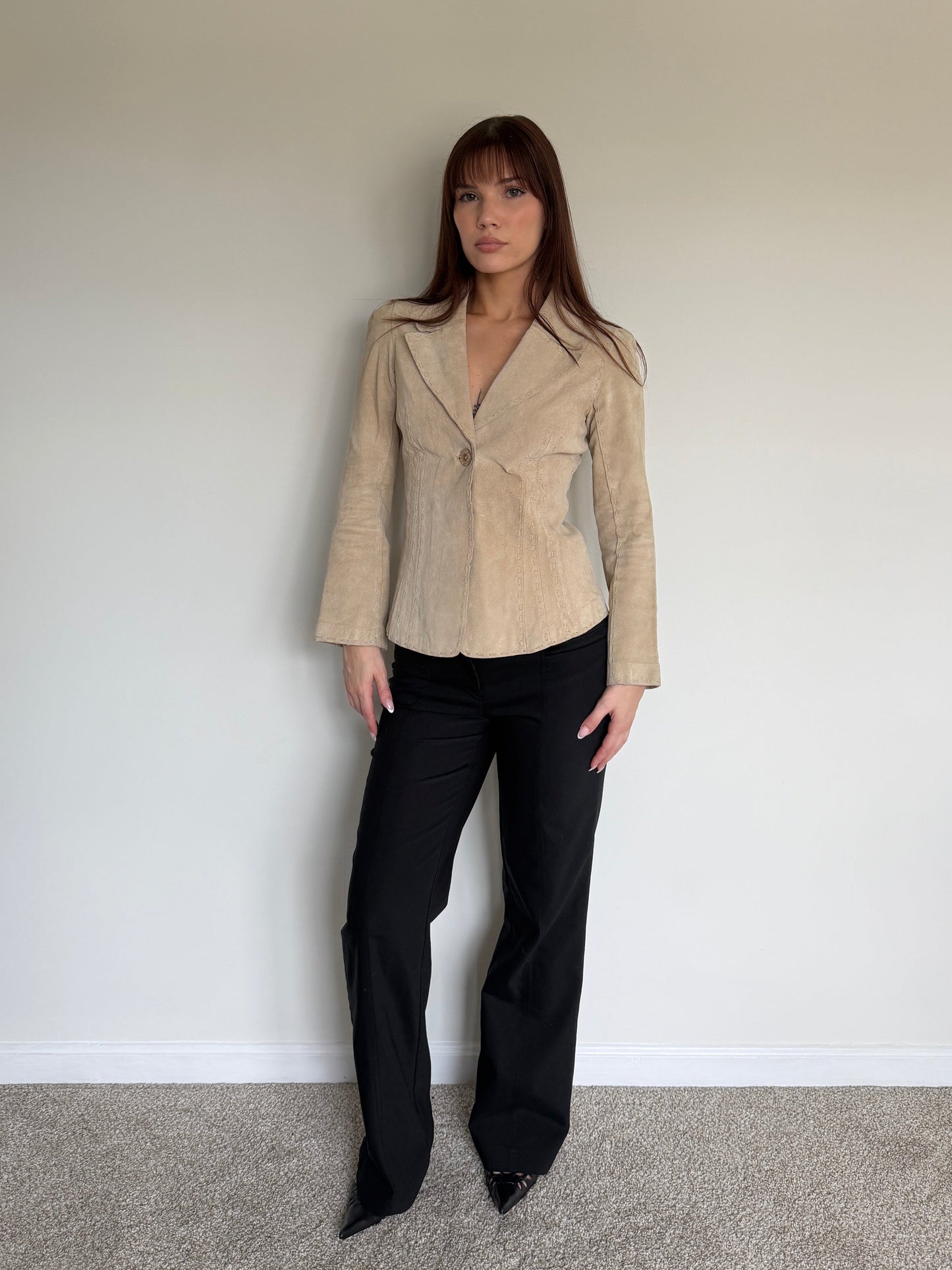 Person wearing a beige blazer and black pants against a plain wall.