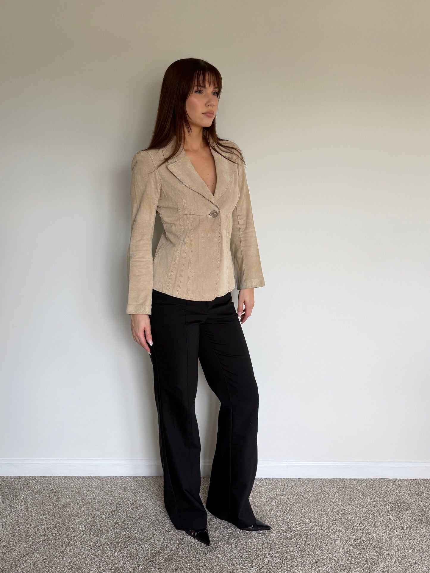 Woman wearing a beige blazer and black pants against a plain wall.