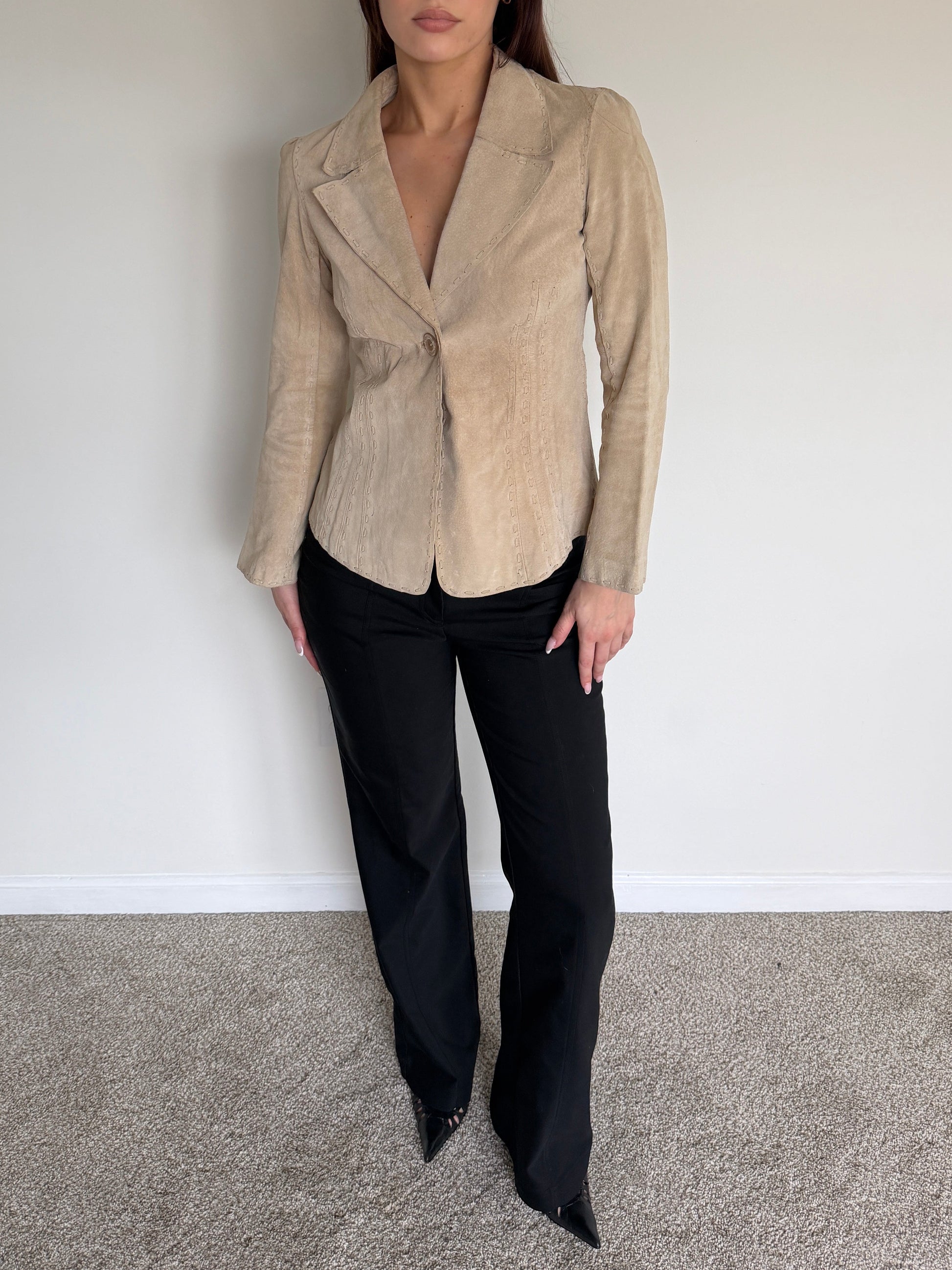 Woman wearing a beige blazer and black pants against a plain white wall.