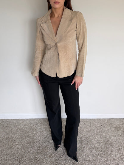 Woman wearing a beige blazer and black pants against a plain white wall.