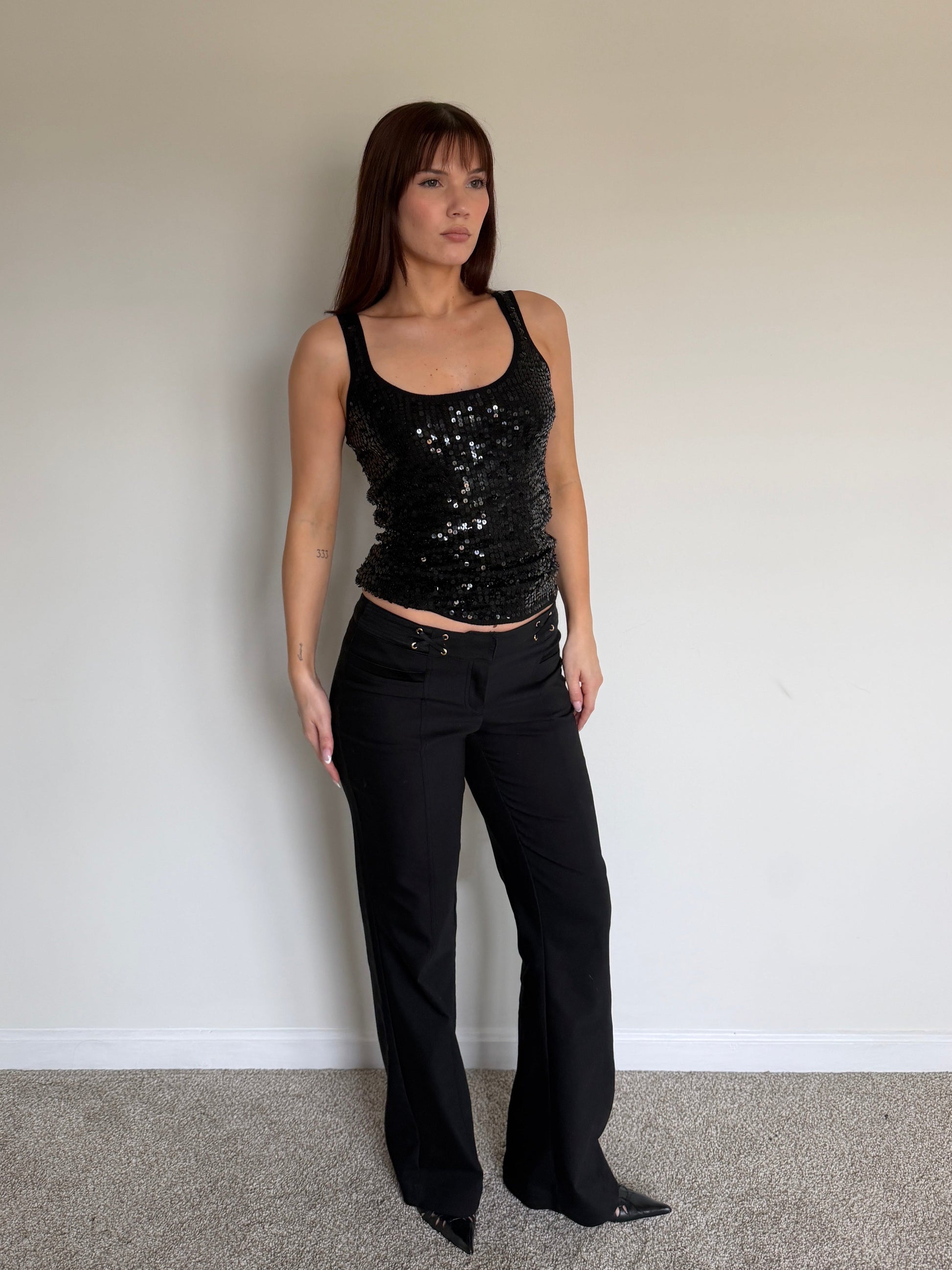 Woman wearing a black sequin top and black pants against a plain wall.