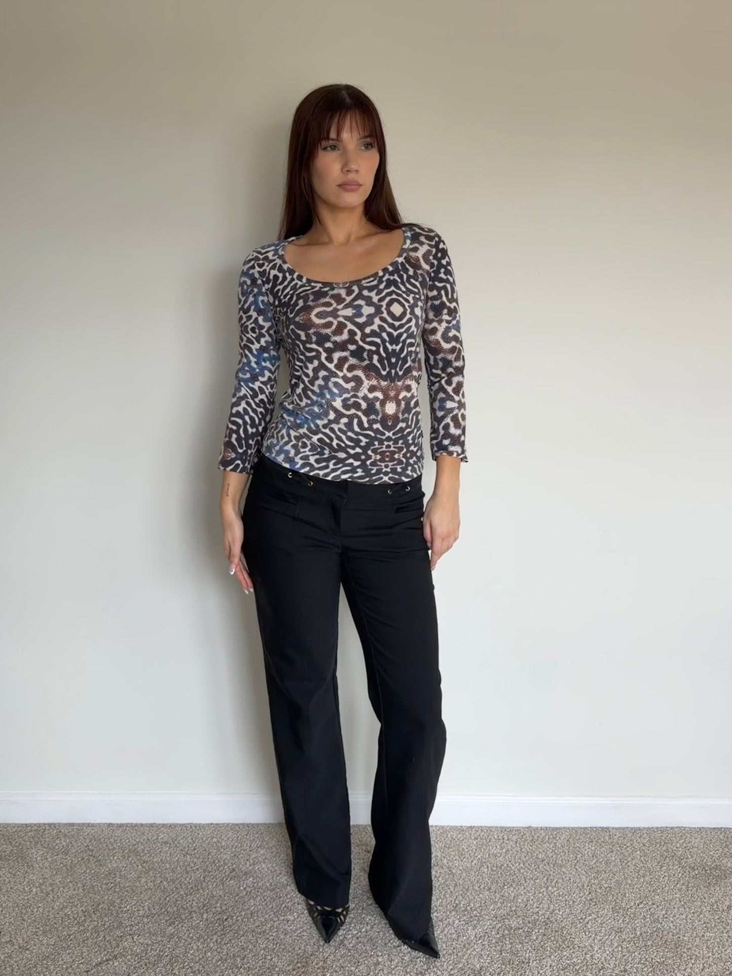 Woman wearing a patterned top and black pants against a plain wall.