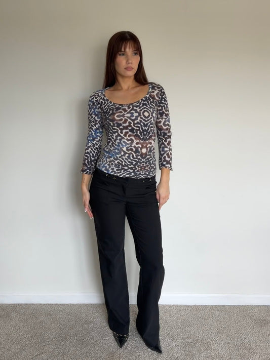 Woman wearing a patterned top and black pants against a plain wall.