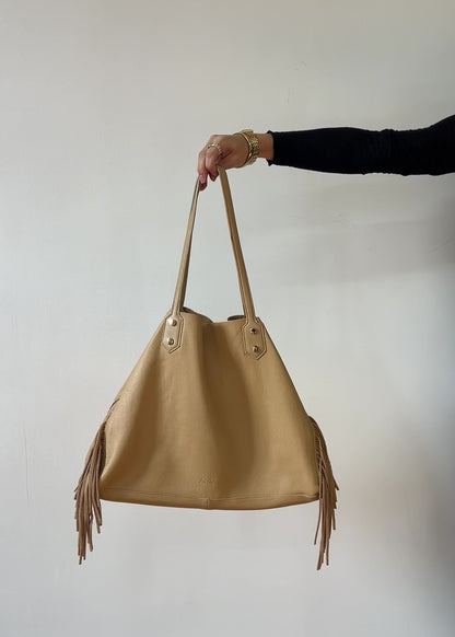 Beige handbag with fringe details held by a person against a plain background